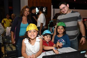 Three children with one with a yellow brain hat and the others with a blue brain hat and a green brain hatreading charts and smilling for the camera.