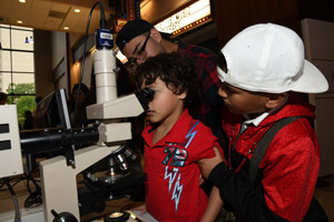 Two children crowded around a microscope with one observing and another anxiously awaiting his turn on the microscope