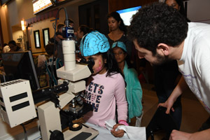  a young girl wearinig a blue brain hat is peering through a microscope at a slice of human brain as a man in a white lab smock explains what she is observing