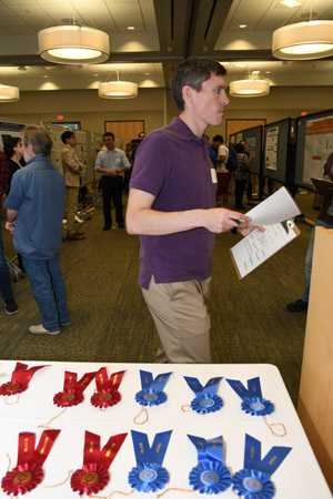 A man in a purple shirt holding a clipboard in front of a table with blue and red winner's ribbons on it.