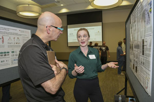 A man with a clipboard watchiing a presentation from a woman presenting her poster.