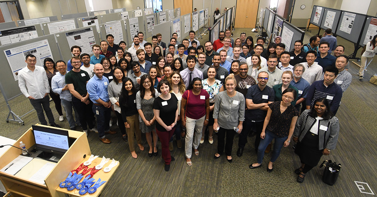 A large group of professors and students standing in front of row of student made posters ready to be judged for their efforts